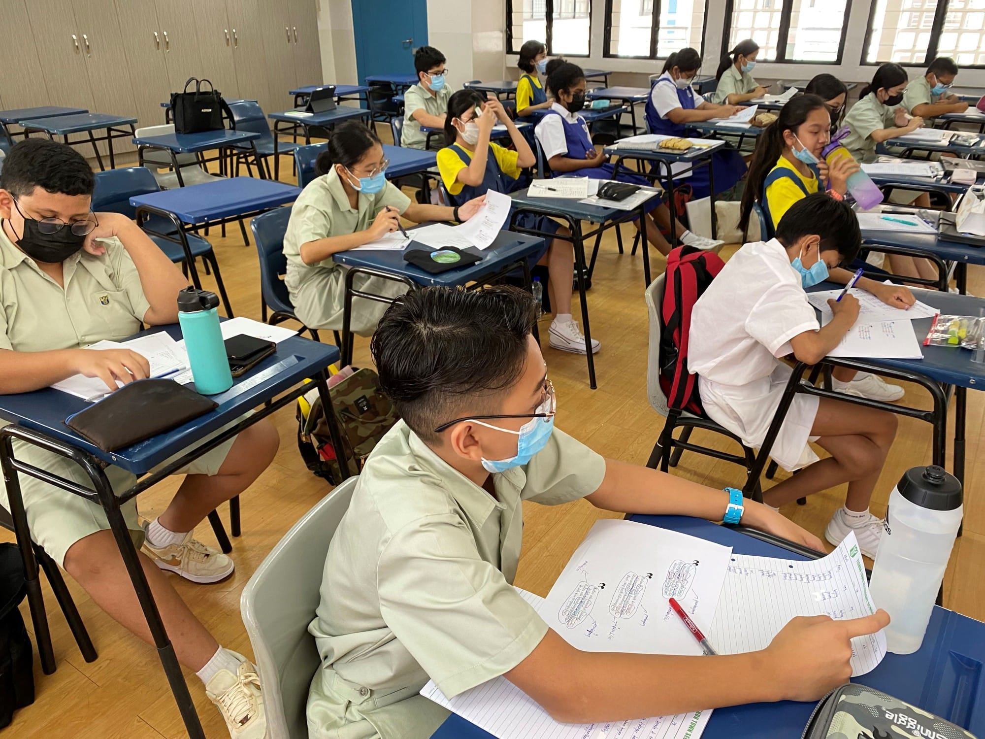 Students in uniforms and face masks sit at blue desks in a classroom, writing on papers.